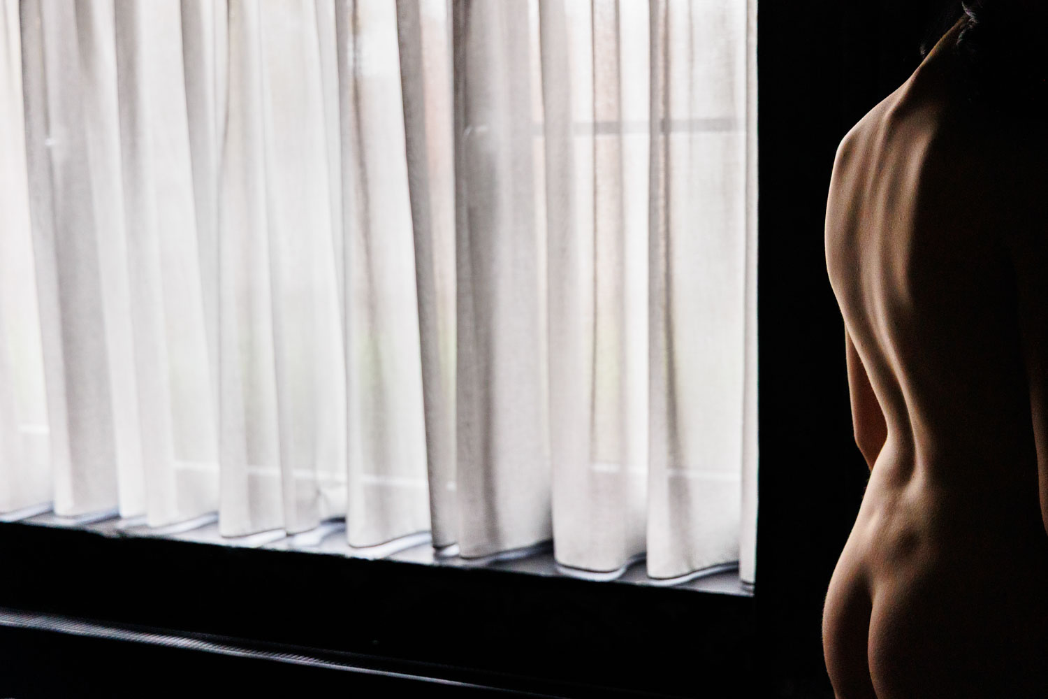 Modelfotografie in huis: naaktstudie van een vrouwenrug met licht en schaduw bij een vitragegordijn